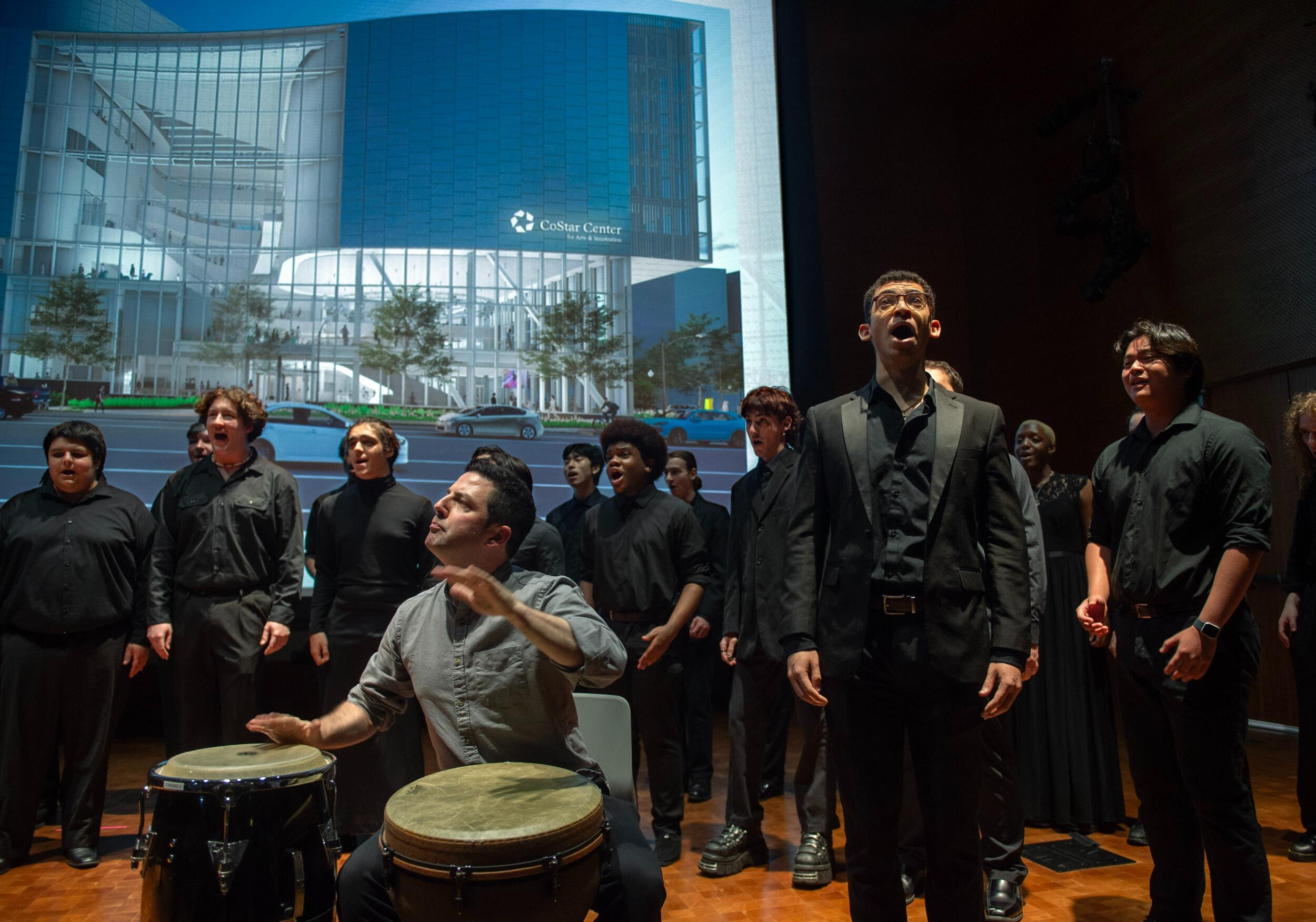 A photo of a group of people wearing all black singing while a man sitting plays the bongo drums in front of them. 