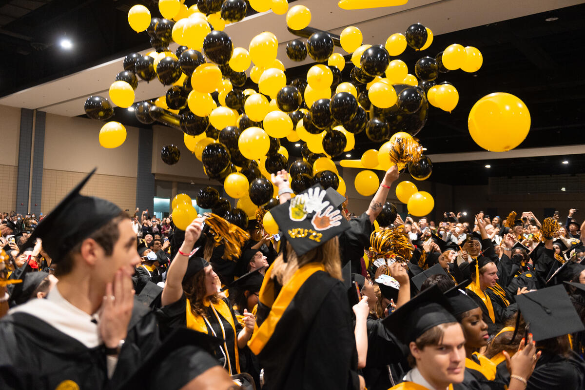 A photo of a crowd of student wearing graduation caps and gowns standing while black and yellow balloons fall from the ceiling. 