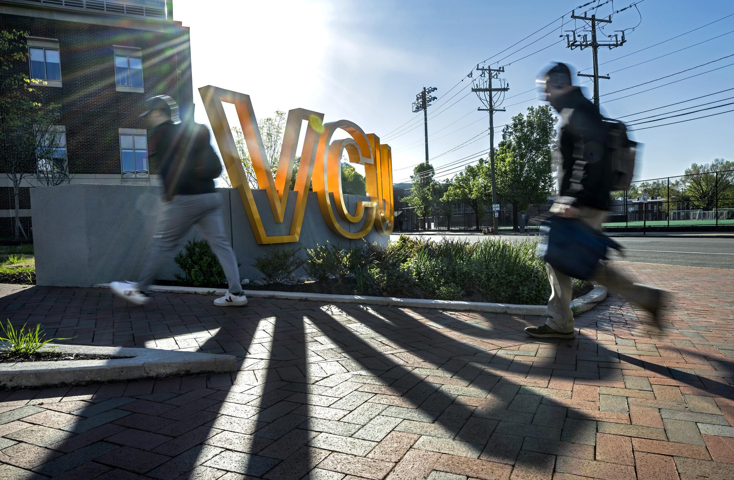 A photo of two people walking on a brick sidewalk. They are passing a sign that spells out \"VCU\" in giant yellow letters. 
