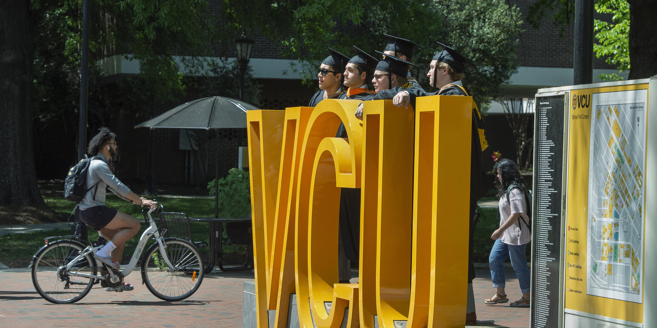 A photo of five people wearing graduation gowns and caps standing behind a sign made up of giant yellow letters that spell out \"VCU.\" To the left of the sign is a man riding his bike past it and to the righ is a woman walking. 