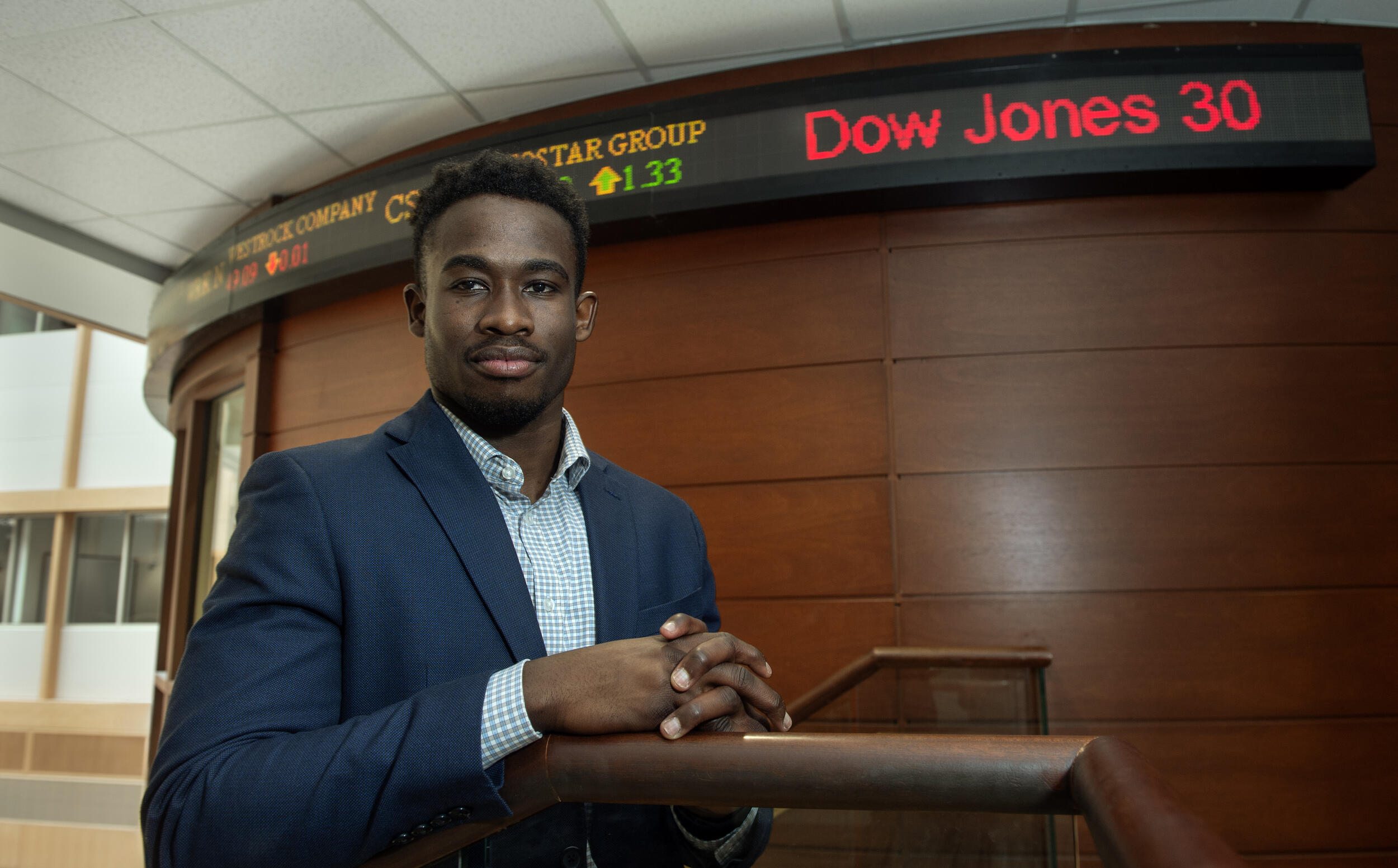 A photo of a man leaning on a railing in front of a curved wall with an electric sign showing stock market updates. 
