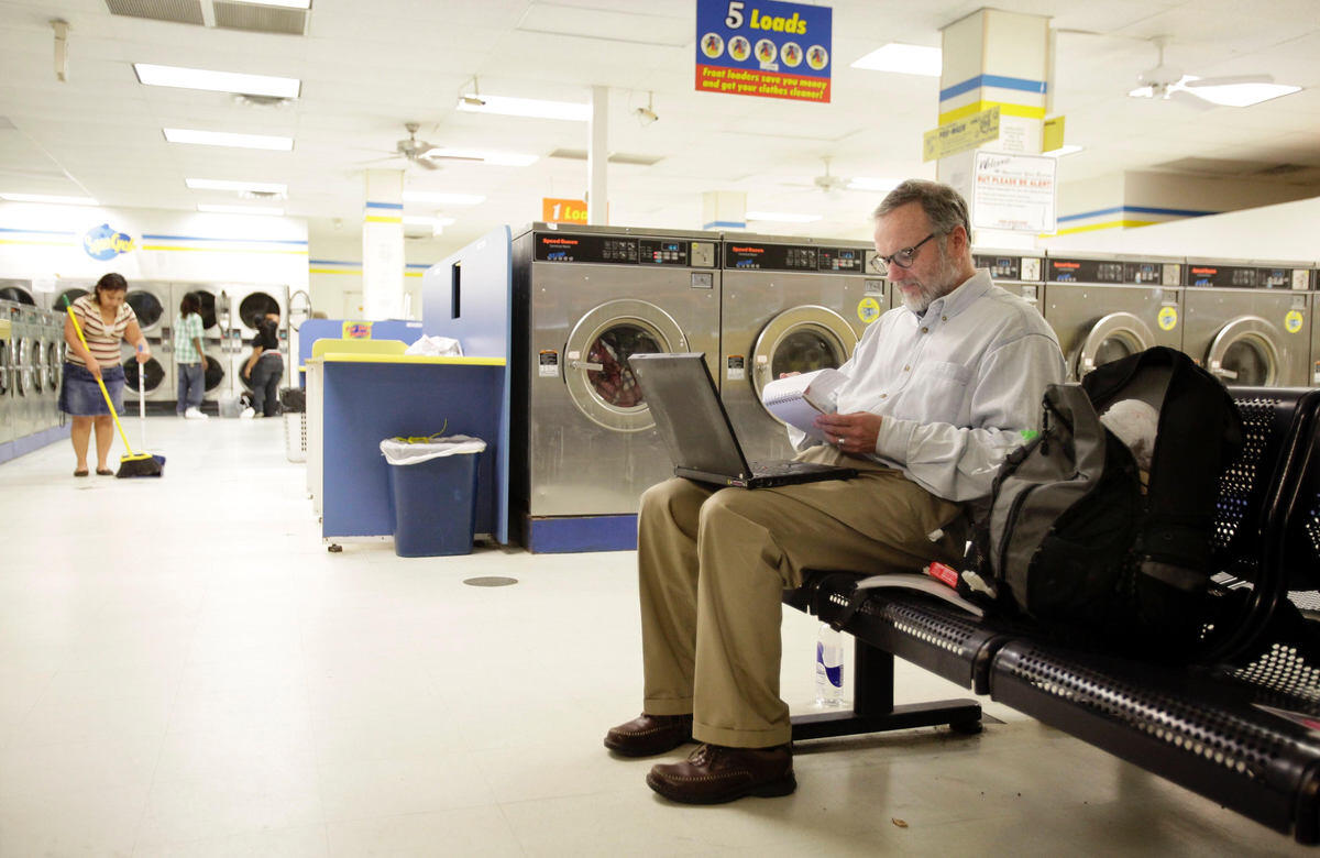A photo of a man sittng on a bench in a laundromat. He has a laptop on his lap and is looking at a notebapd. He has a black backpack next to him. In the background a woman is sweeping the floor and two other people are standng in front of a row of washers. 