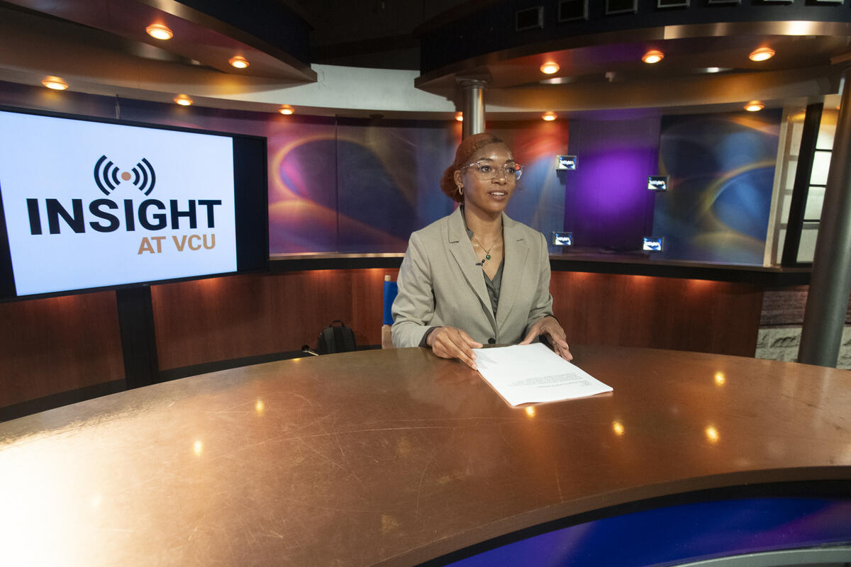 A photo of a woman wearing glasses sitting at a news anchor desk with a sheet of paper in front of her. Behind the woman is a monitor that says \"INSIGHT AT VCU\" in yellow and black letters. 