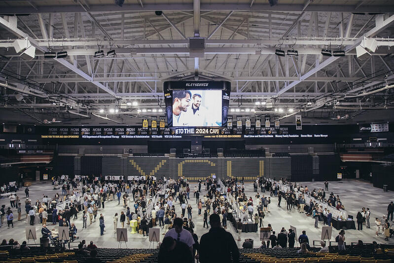 A phot of the basket ball court in the Siegel Center's basketball court floor full of tables with posters on them. The room is full of people around the booths, and a few people are sitting in the bleachers. 