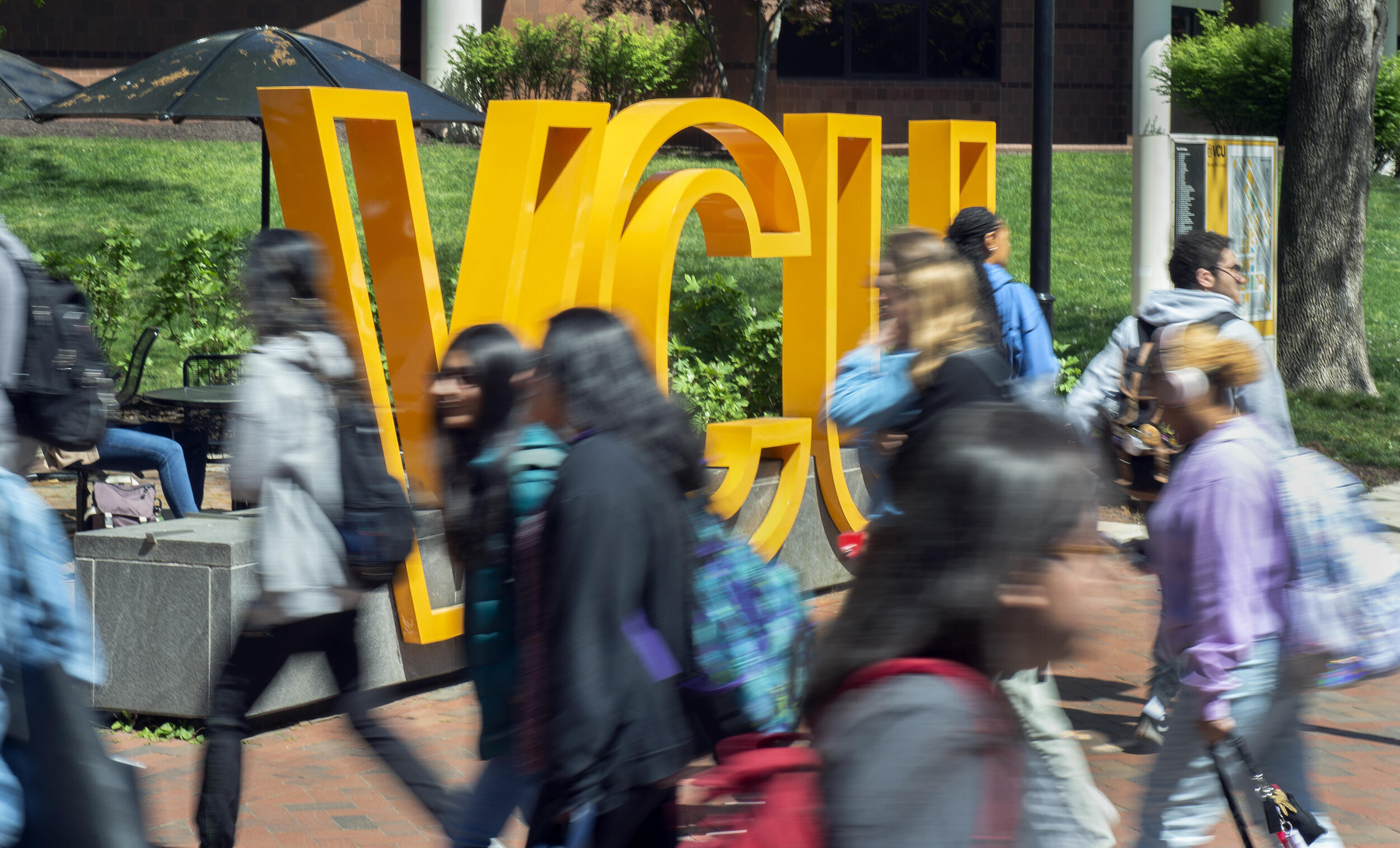 A photo of people walking on a sidewalk past a sign with large yellow letters that spell out \"VCU\" 