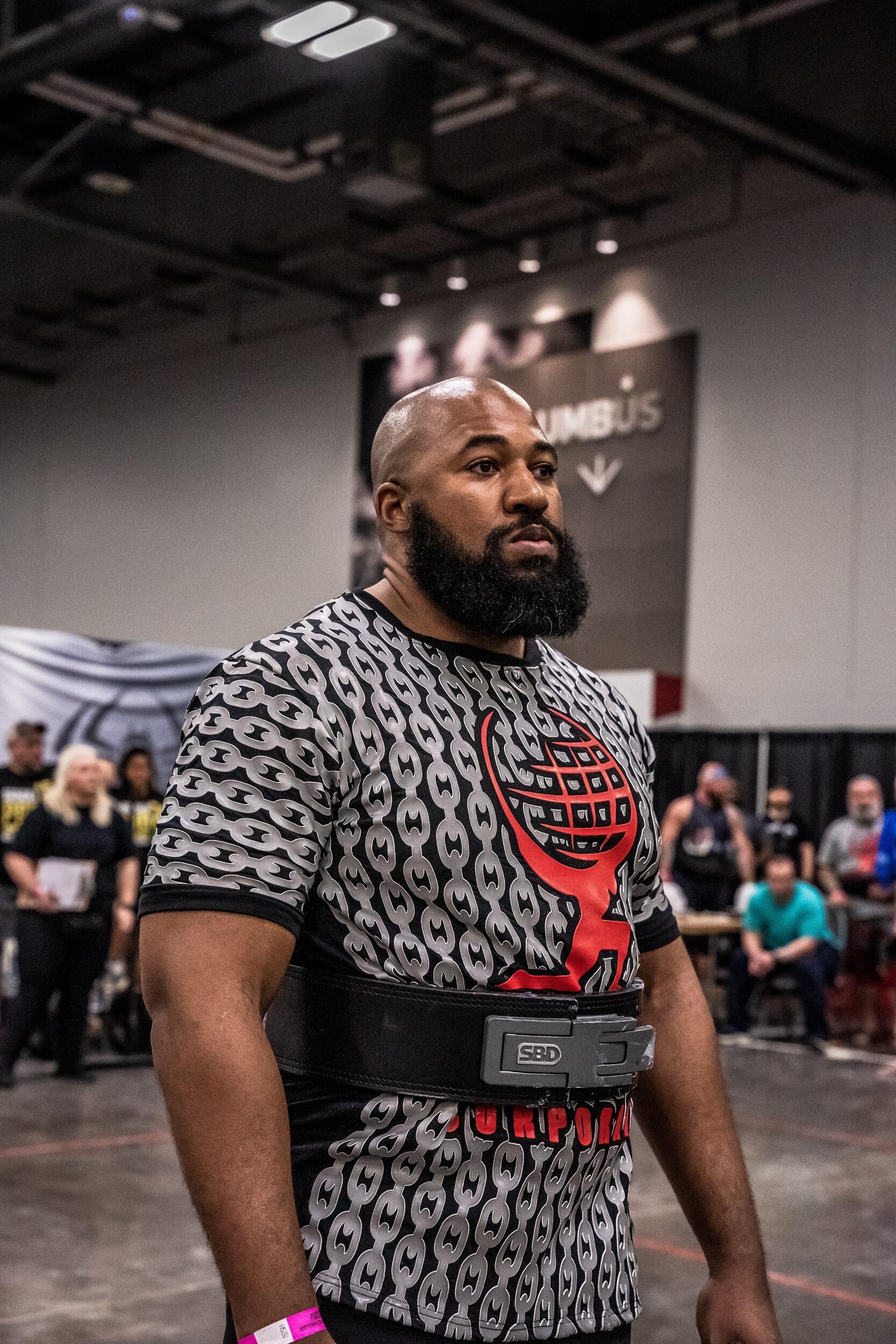 A photo of a man from the waist up wearing a t-shirt and a weight lifting belt. 