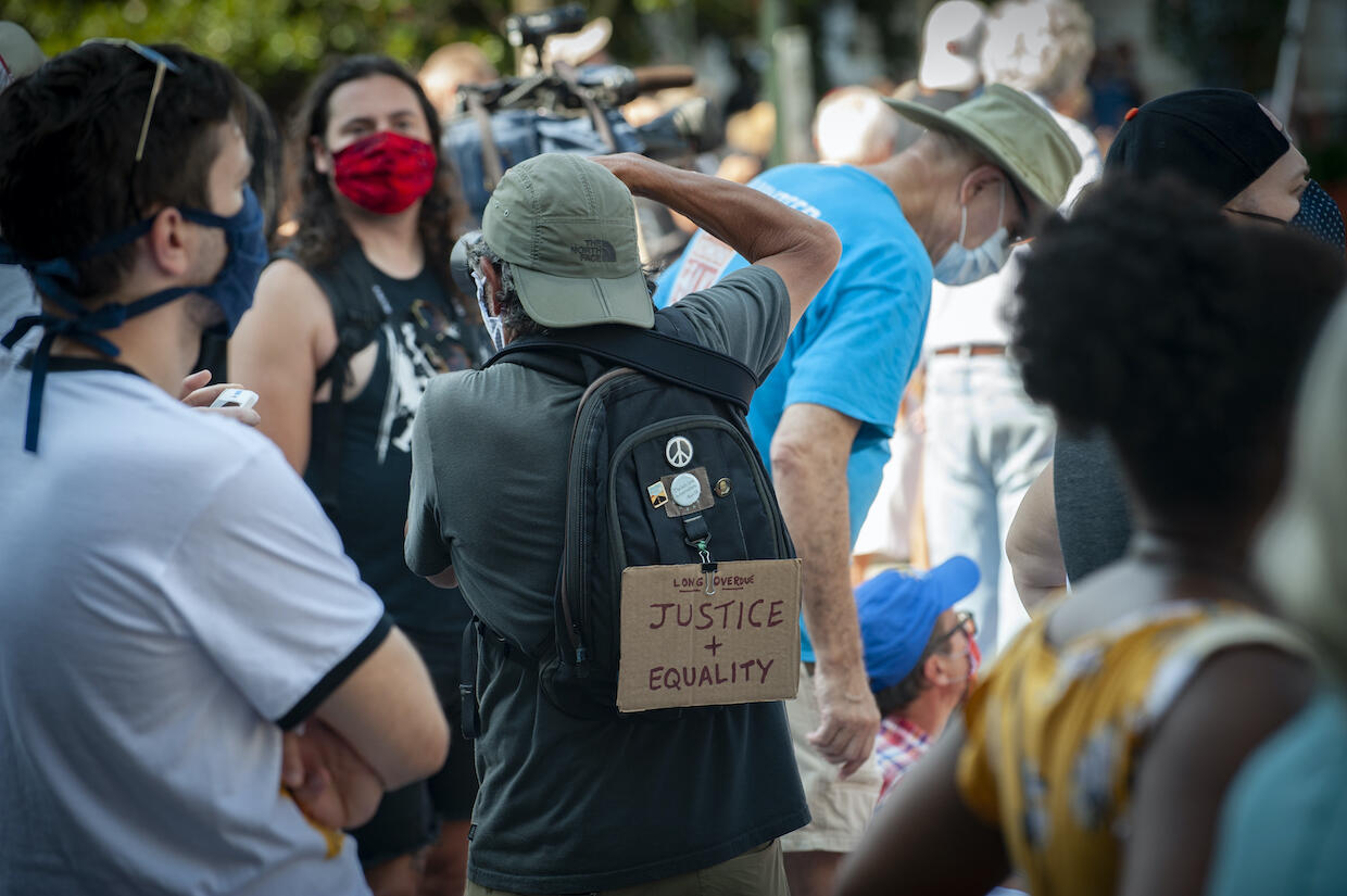 A group of people standing. A person wearing a backpack with a sign stating \"Long Overdue. Justice + Equality\" takes a picture.