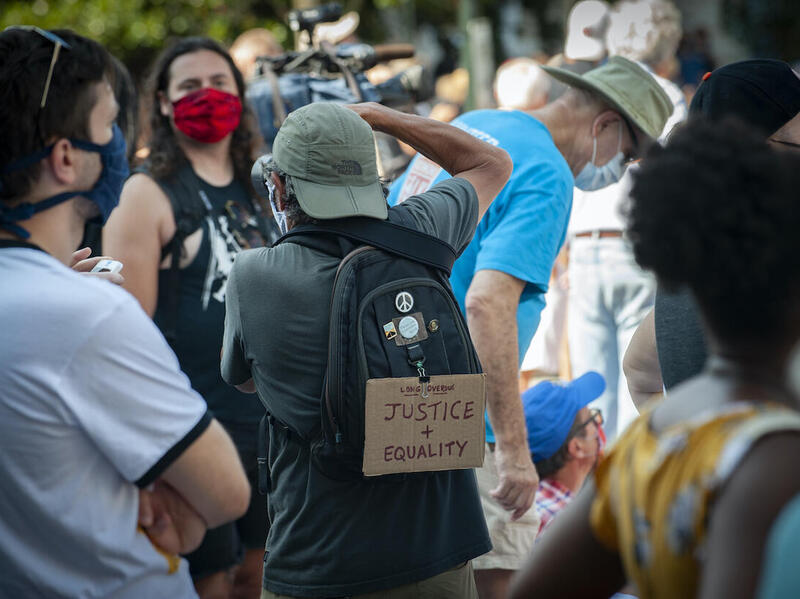 A group of people standing. A person wearing a backpack with a sign stating \"Long Overdue. Justice + Equality\" takes a picture.