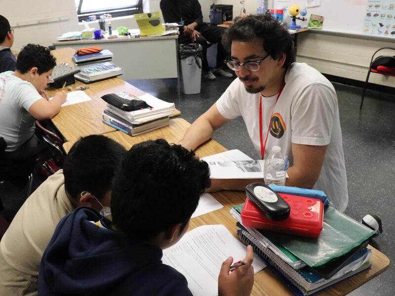A photo of a a man sitting at a desk with two childre. Next to them are two other children sitting at another desk. 