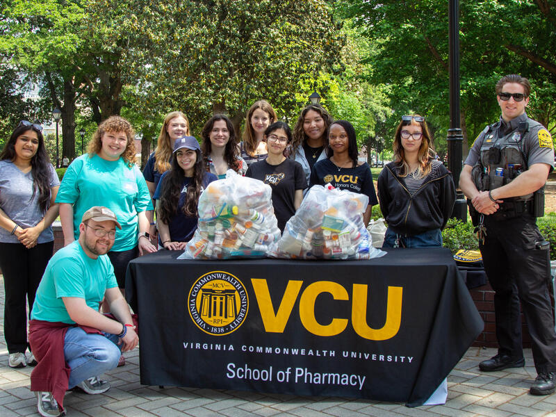 A group photo of 10 people standing around a table and one person kneeling next to it. The table has two large clear, plastic, trash bags filled with perscription pill bottles. The table has a table cloth that says \"VCU\" in large yellow letters. In smaller white letters the table cloth says \"V I R G I N I A C O M M O N W E A L T H U N I V E R S I TY\" and \"School of Pharmacy>' 