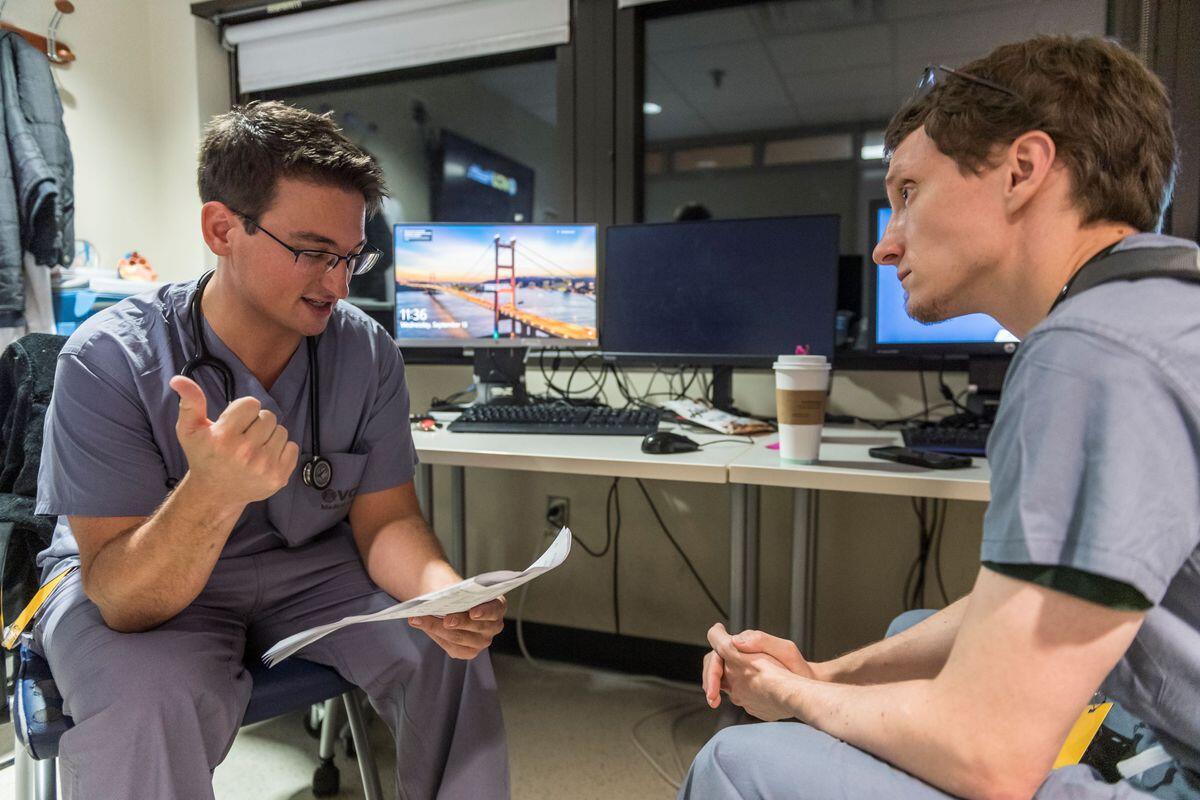 A photo of two men wearing scrubs sitting in chairs facing each other and talking. 