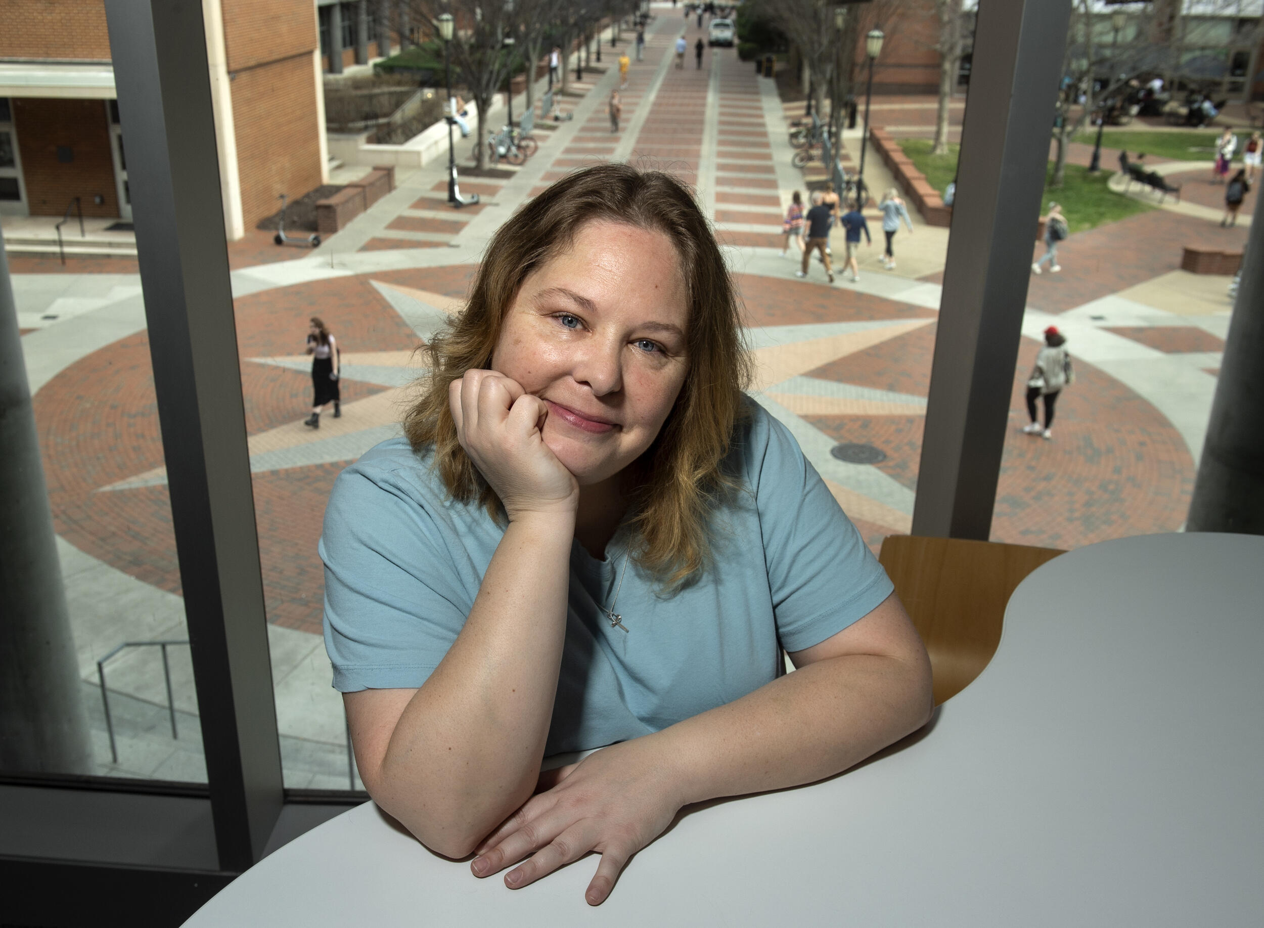 A woman sitting at a table with her left arm resting on it and her head sitting in her right hand. She is sitting in front of a window that faces the compass plaza. 