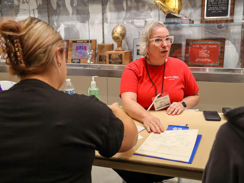 A woman in a red T-shirt sits at a table with a piece of paper in front of her. Another woman is the foreground with her back to the camera.