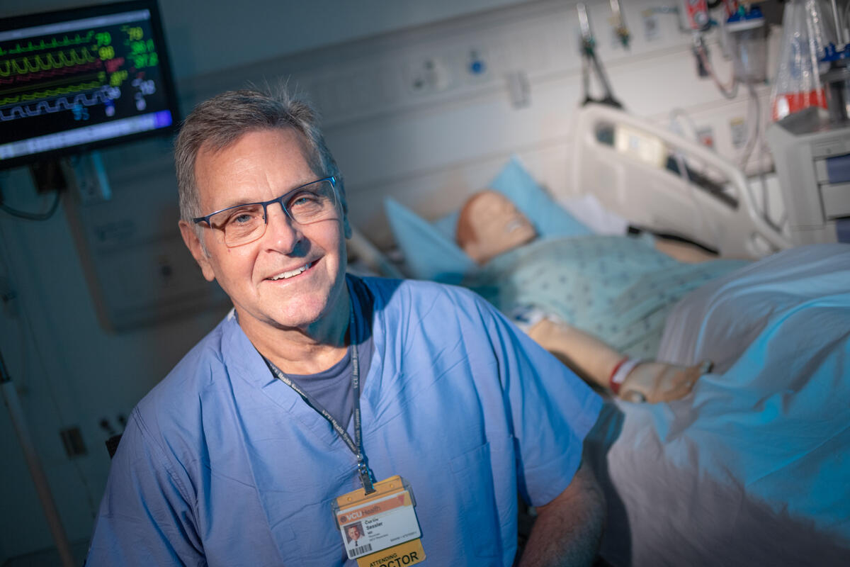 A photo of a man standing in front of a hospital bed that has a dummy laying in it. 