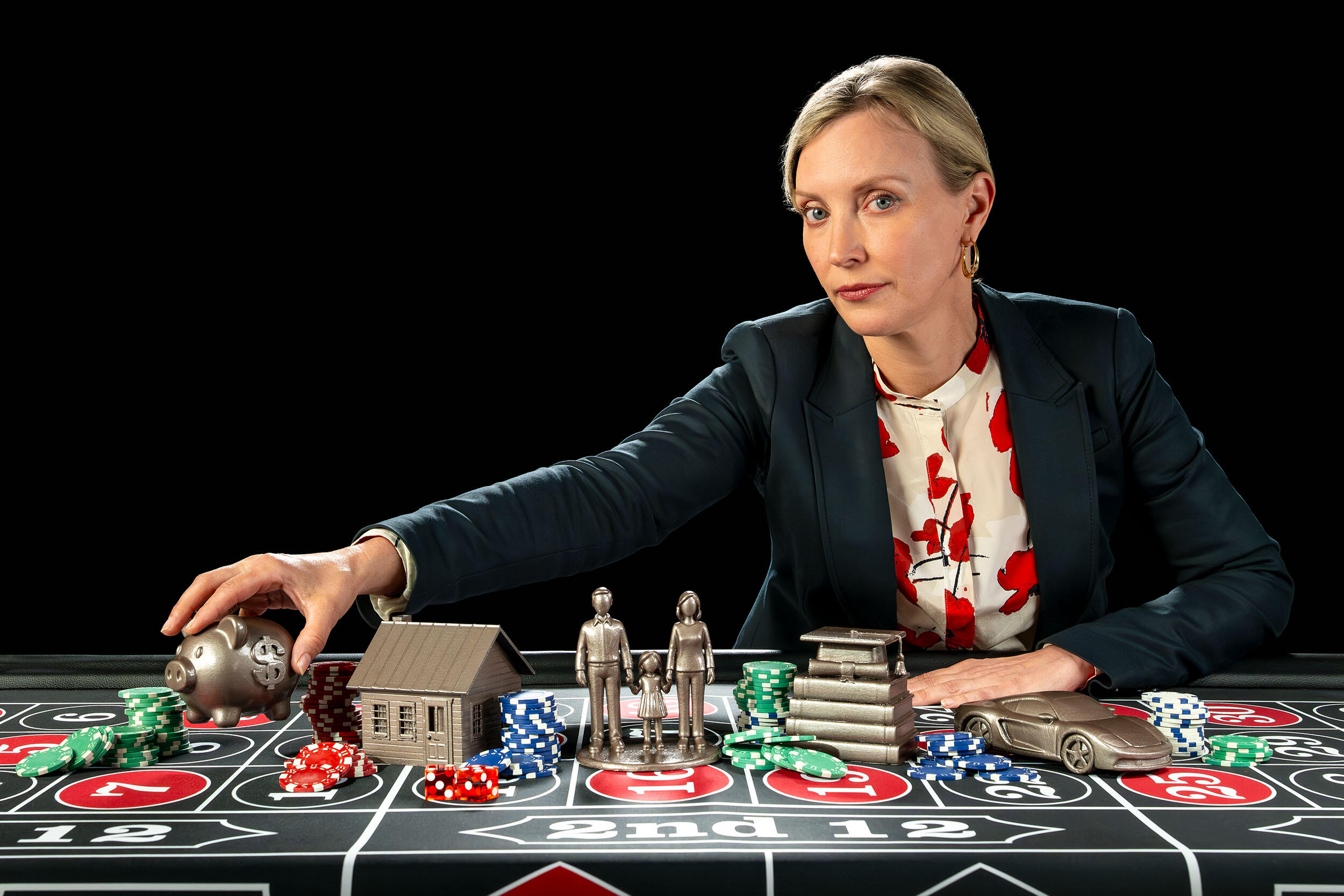 Carolyn Hawley reaches across a gambling table filled with poker chips and small statues of a house, people, a stack of books and a car; she holds a piggy bank. 