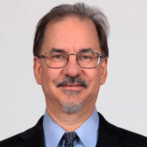 A portrait of a man wearing glasses, a light blue shirt, a dark blue tie with light blue dots, and a black sportscoat. 