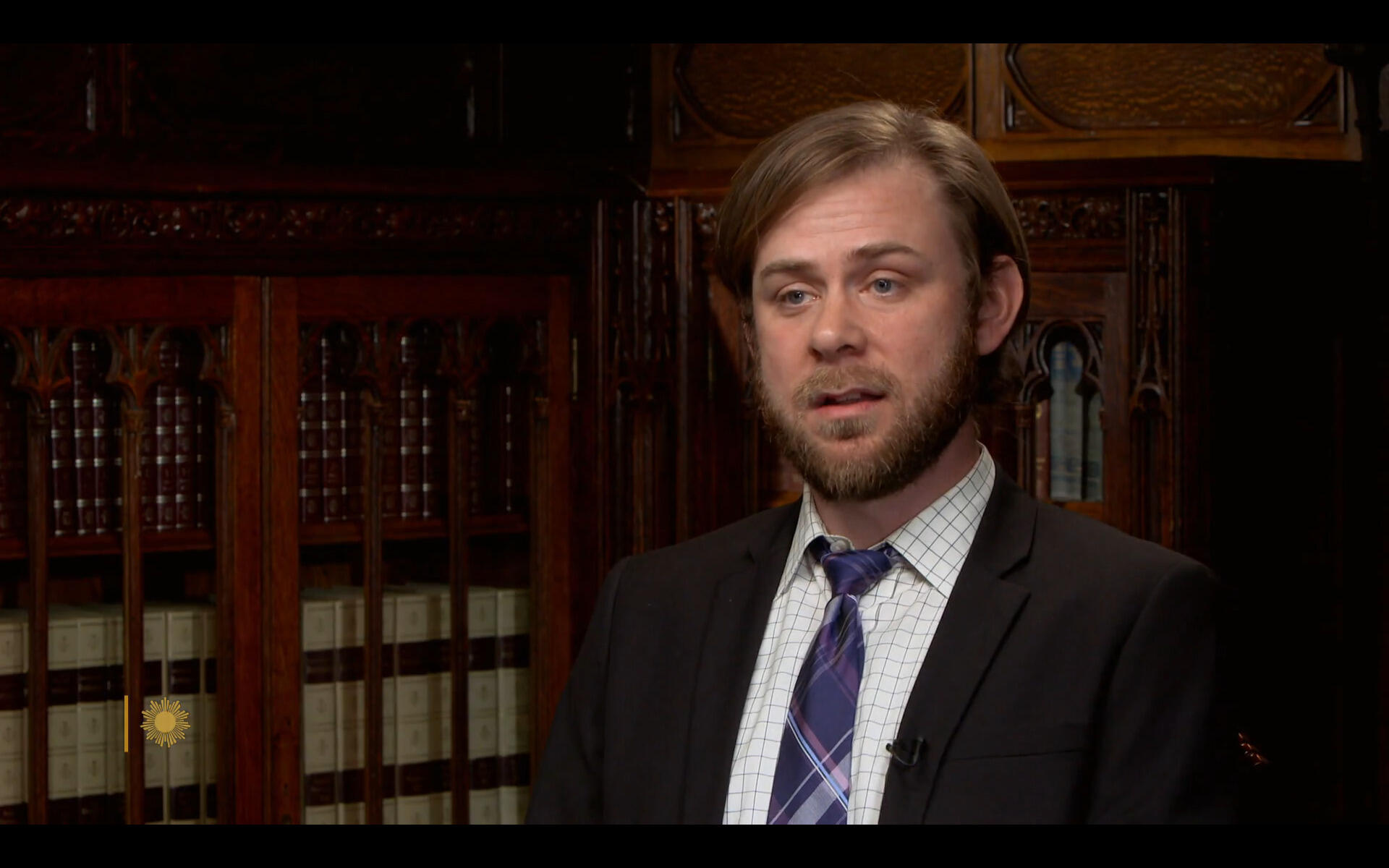 A man wearing a sportscoat, dress shirt, and tie sitting in front of a bookshelf 