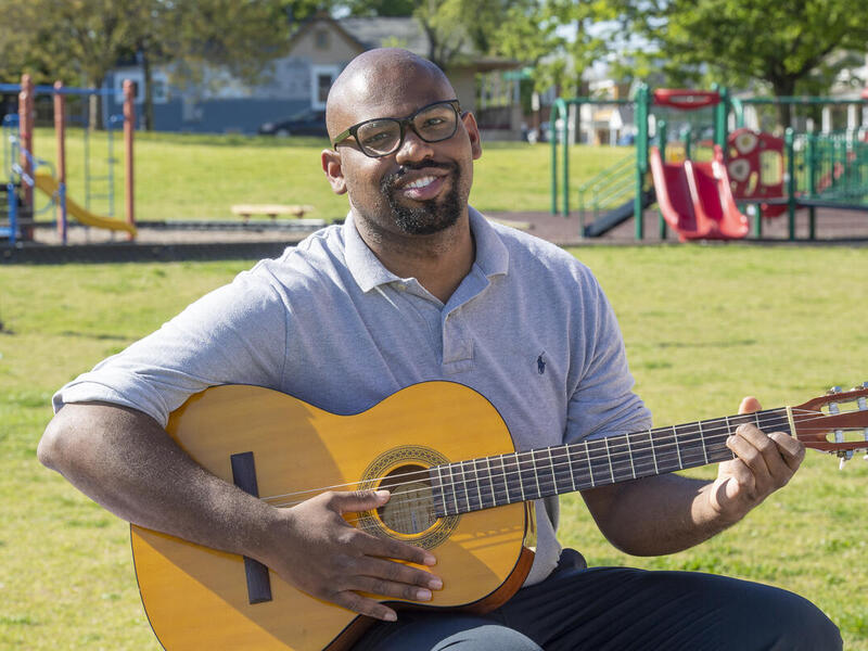 A man sitting in the grass holding an acoustic guitar. Behind him is a playground. 