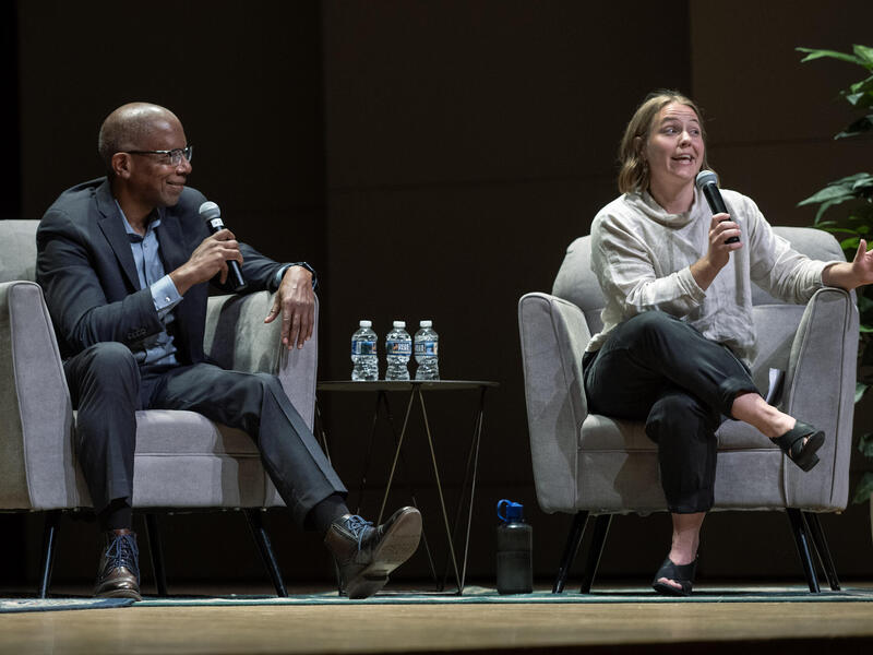 A photo of a man and woman sitting in two gray arm chairs. Each person is holding a microphone. The man is on the left watching the woman on the right speak. 
