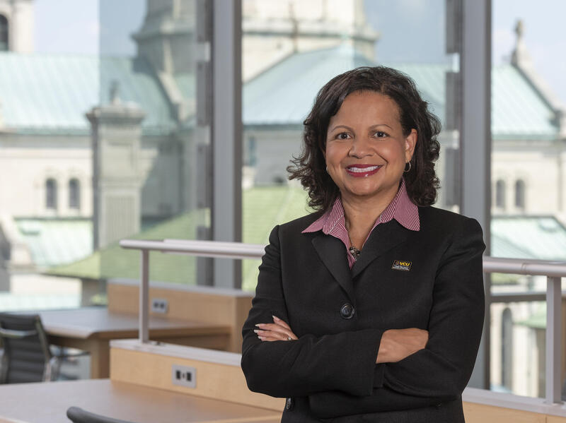 A photo of a woman from the waist up. She is standing in front of a window with her arms crossed against her chest. 