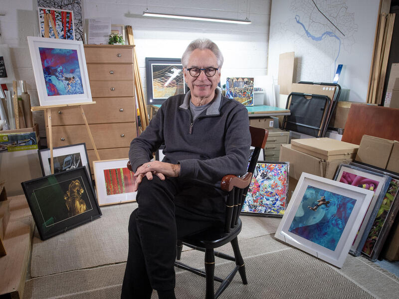A photo of a man sitting in a chair in the middle of a room. Around him are several paintings sitting against boxex and on an easle. 