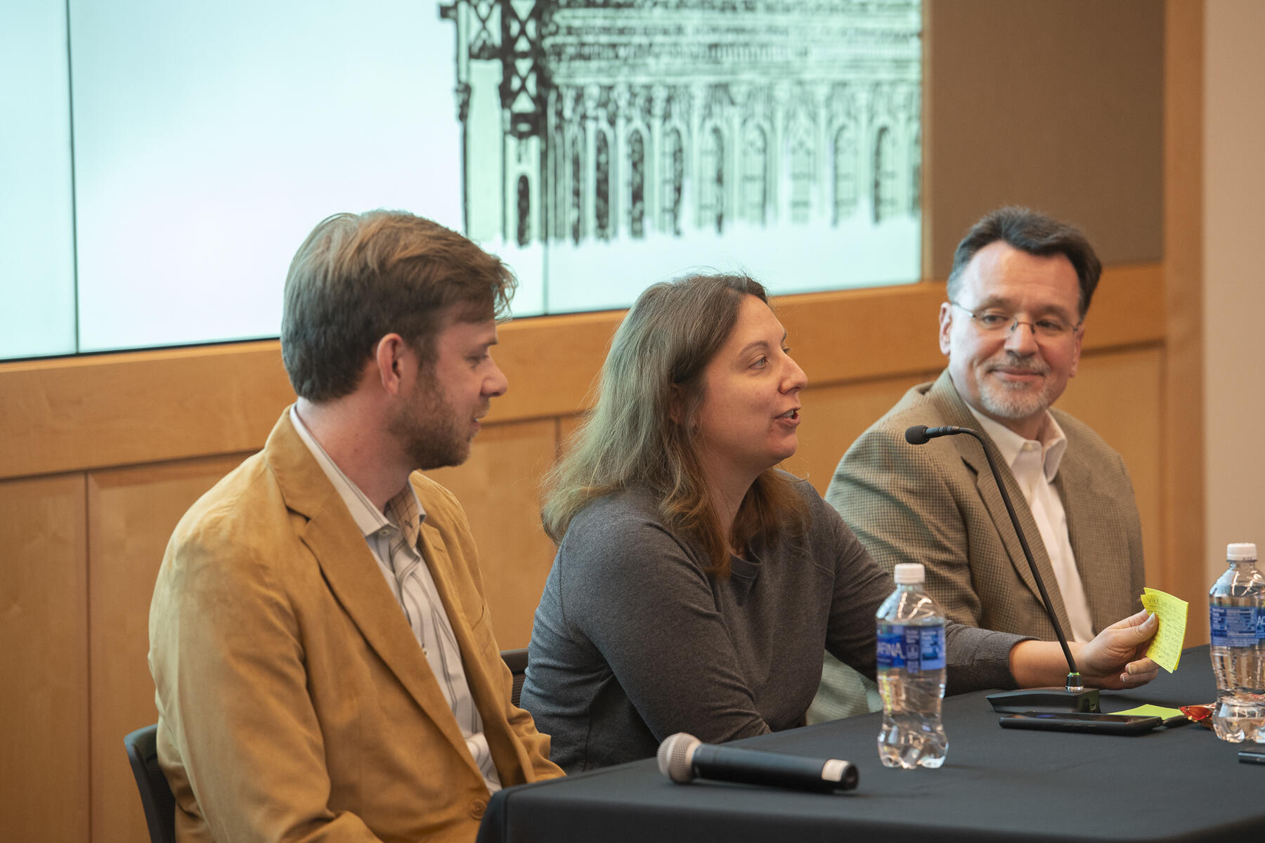 A photo of two men sitting on either side of a woman at a table. The men are both watching the woman speak. 