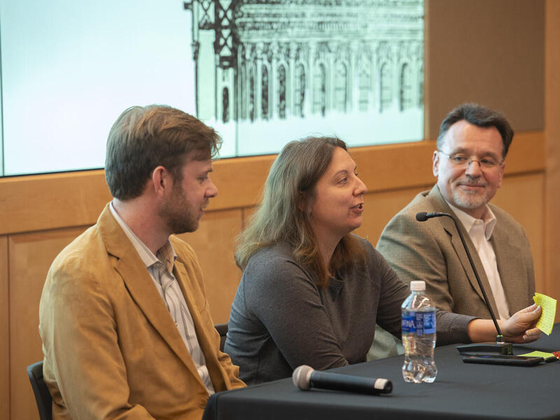 A photo of two men sitting on either side of a woman at a table. The men are both watching the woman speak. 