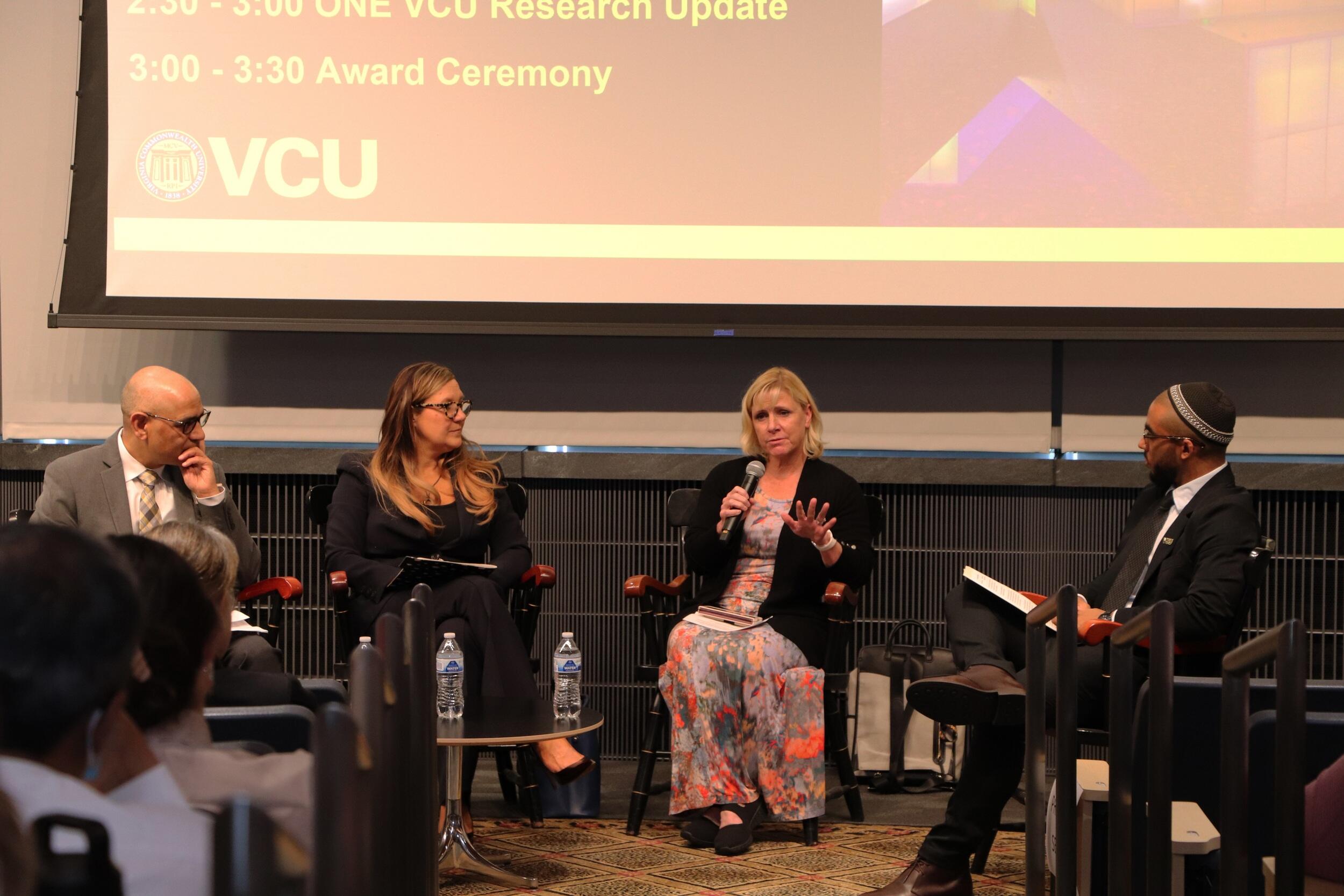 A photo of four people at the front of a room spekaing in front of a projector. 