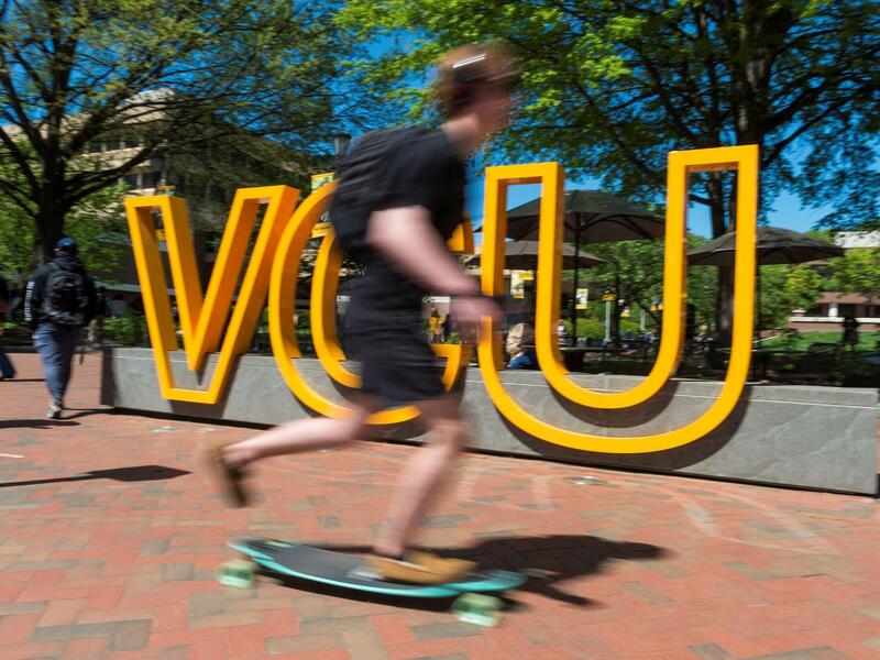 A person rides a skateboard in front of large VCU letters on a sidewalk.