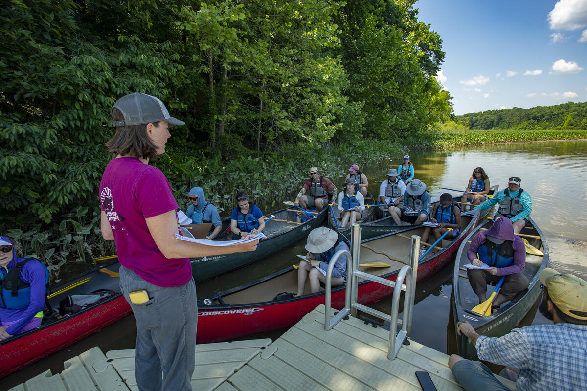 A woman standing on a dock talking to a group of people sitting in canoes on the river 
