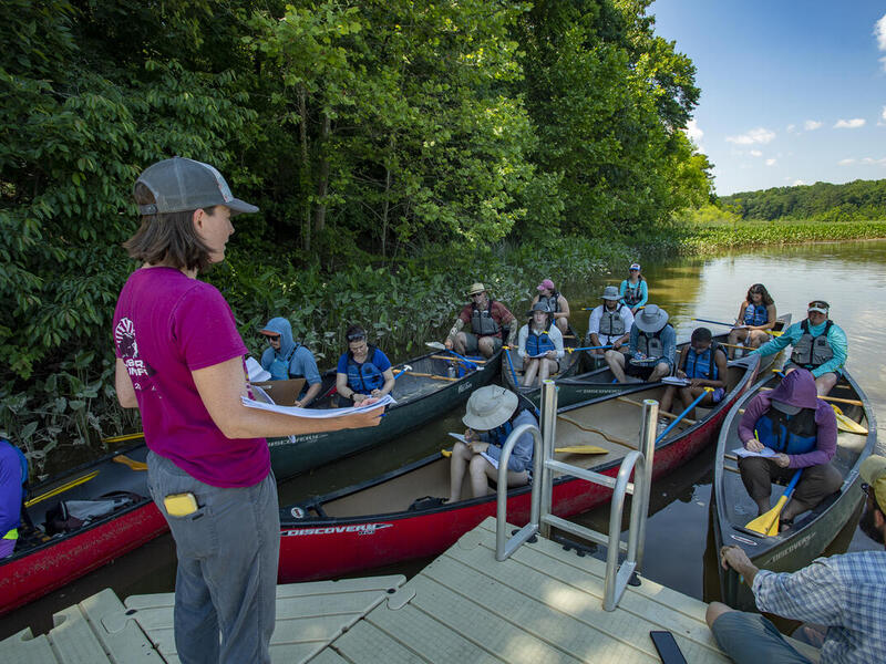A woman standing on a dock talking to a group of people sitting in canoes on the river 