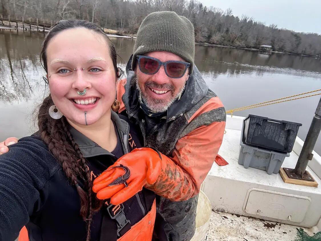 A photo ofa a man and woman smiling on a boat. The man is holding a small fish in his hand. 