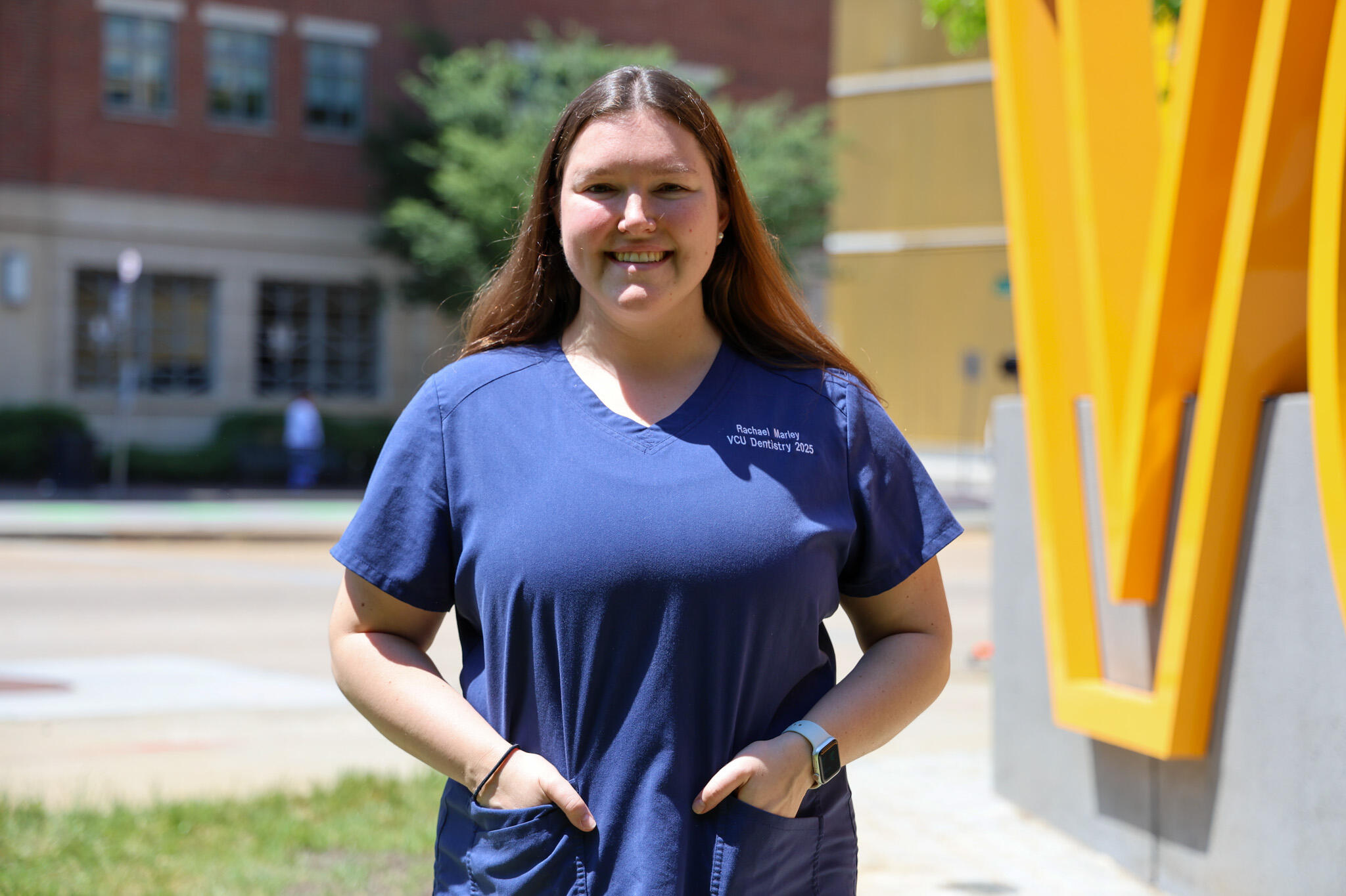 A photo of a woman from the waist up. She is wearintg blue dental scrubs and has her hands in the pockets of the shirt. 