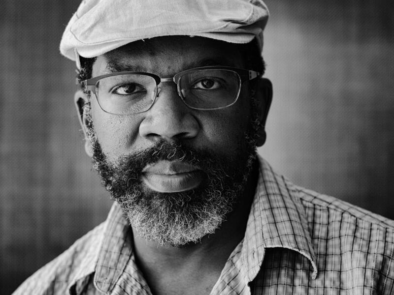 A black and white photo of a man with a short beard wearing a hat and glasses. 