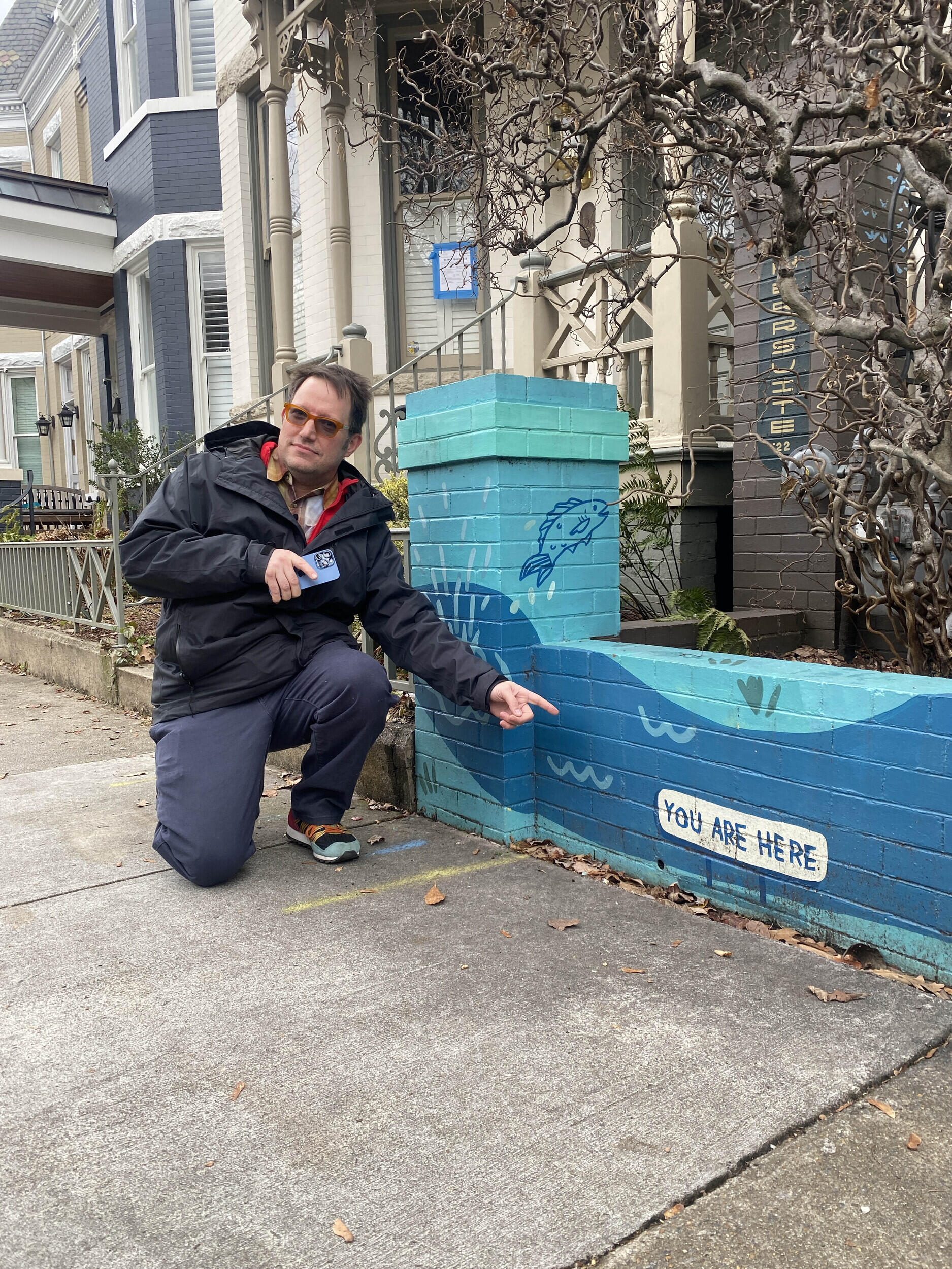 A photo of a man kneeling next to a brick fence painted blue. On the fence is a white bubble with blue text that reads \"you are here.\" The main is pointing at the text with his right hand. 