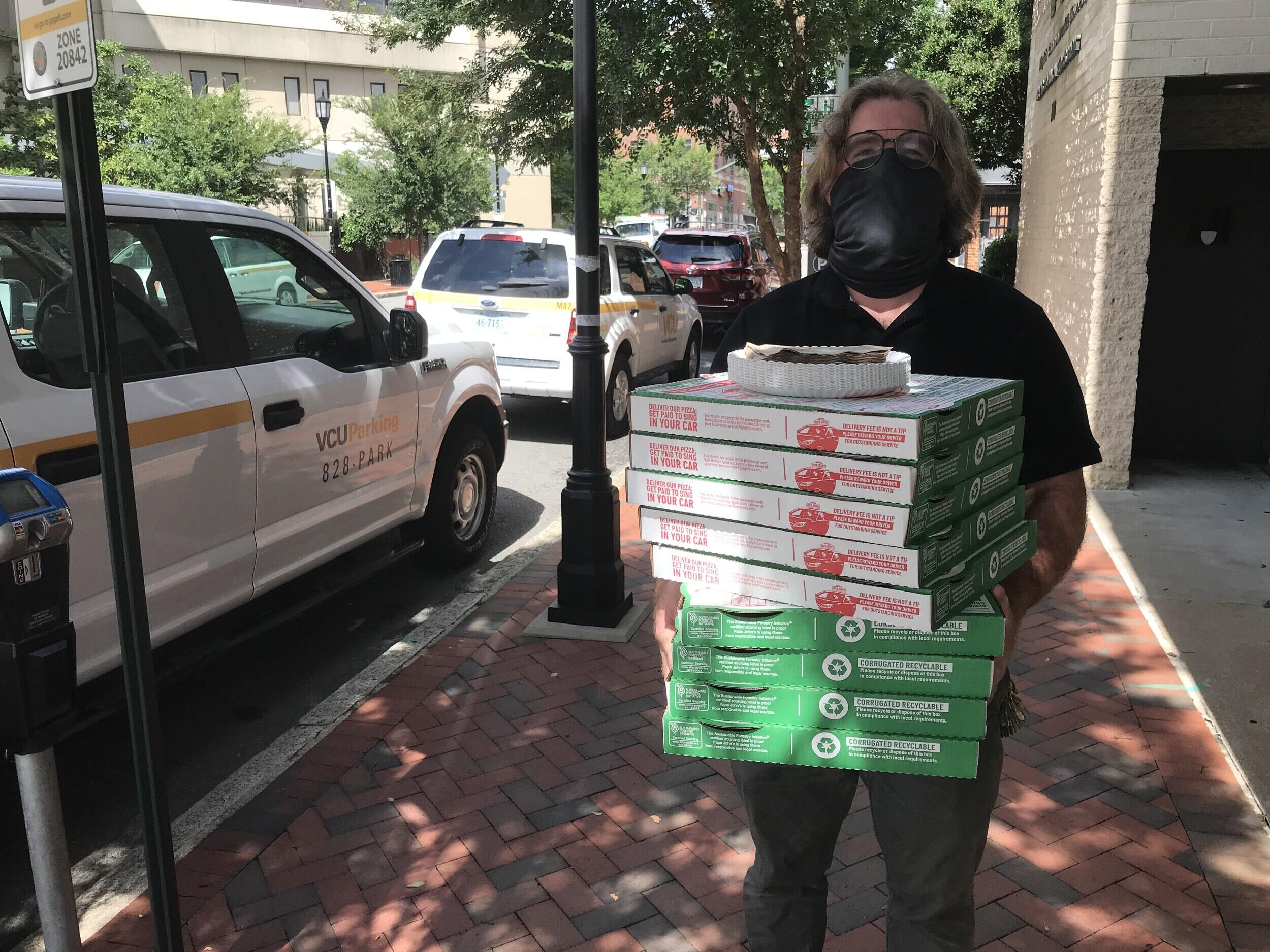 A person wearing a mask carries boxes of pizza, paper plates, and napkins.