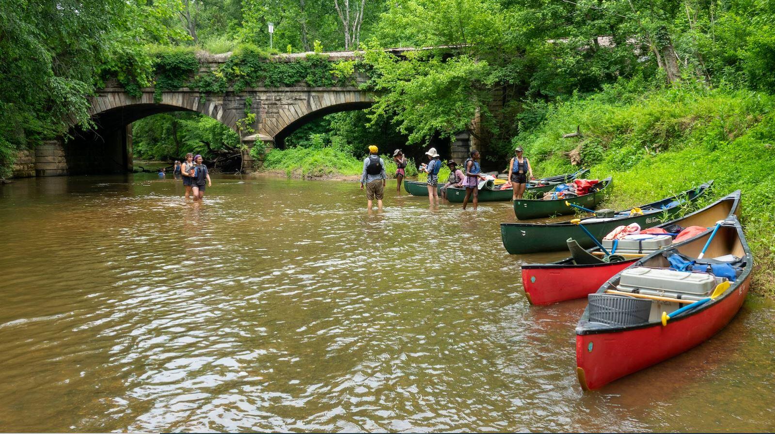 Boats on the shore of a river. People are standing further down the river in front of a bridge 