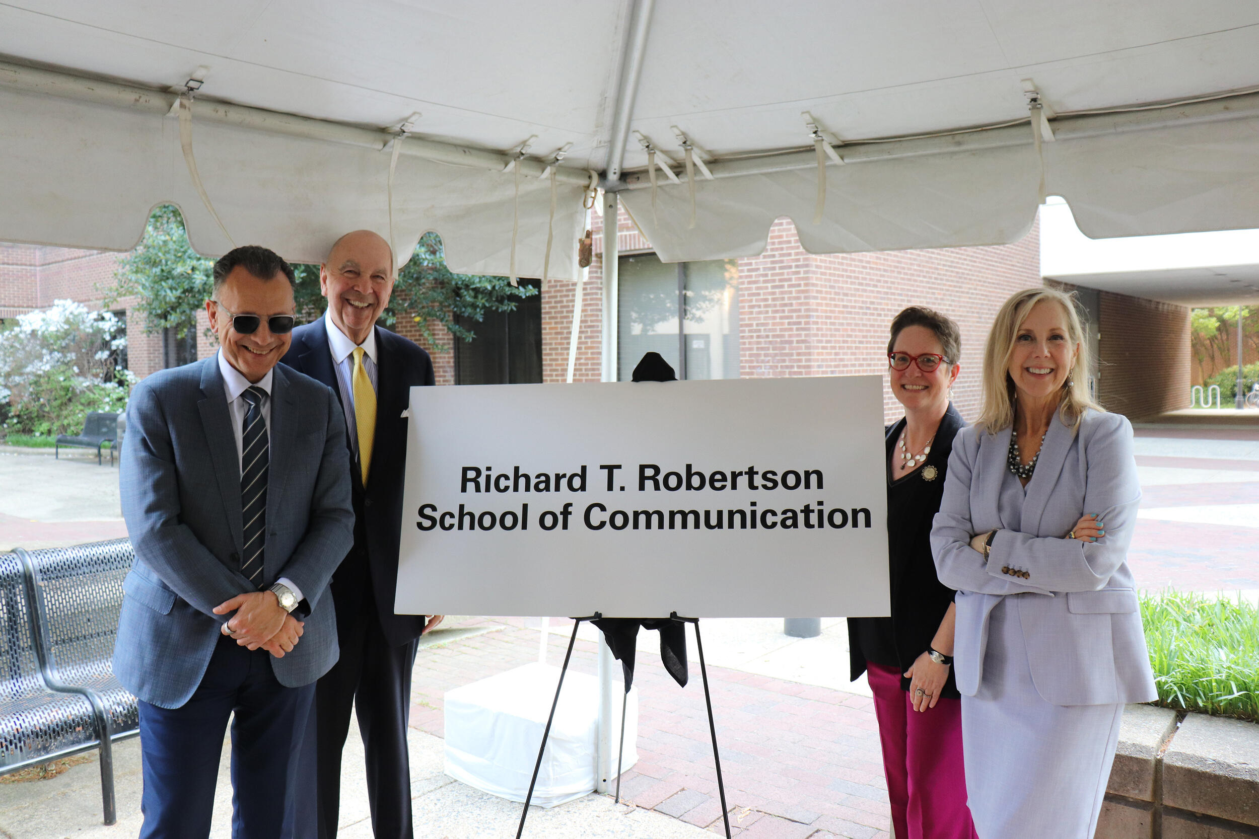 A photo of two men standing to the left and two women standing to the right of a white sign. The sign says \"Richard T. Robertson School of Communication\" in black letters. 