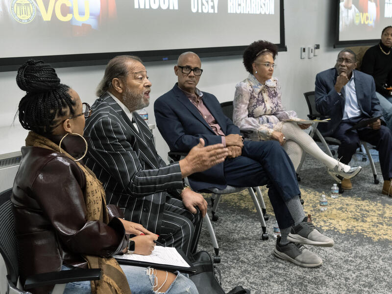 A photo of give people sitting at the front of a room with a projector behind them. The second person to the right is speaking and everyone is looking at him. Another person is standing to the right of the five people sitting, looking over at the man talking. 
