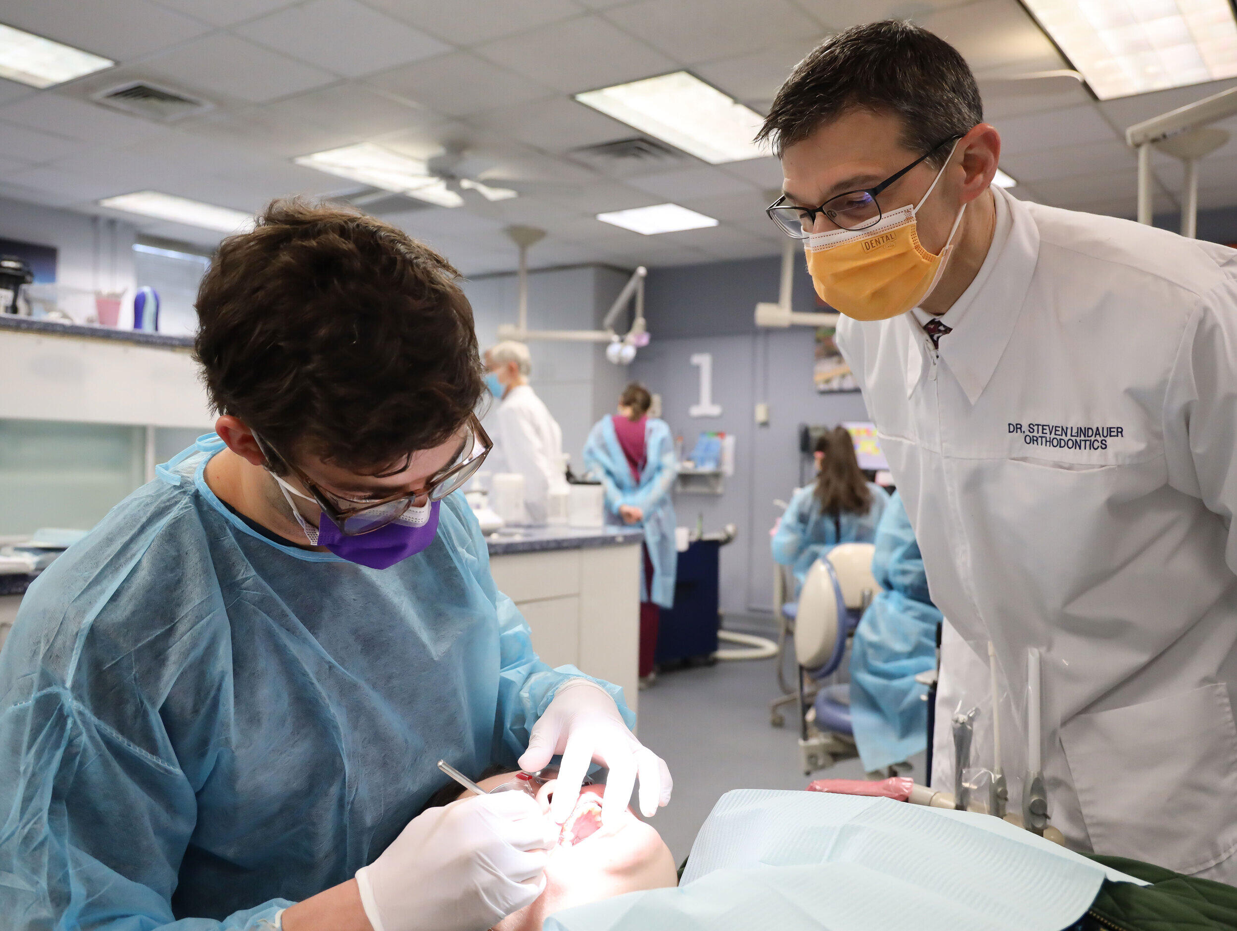 A photo of a dental professor watching a dental student clean a patient's teeth. 