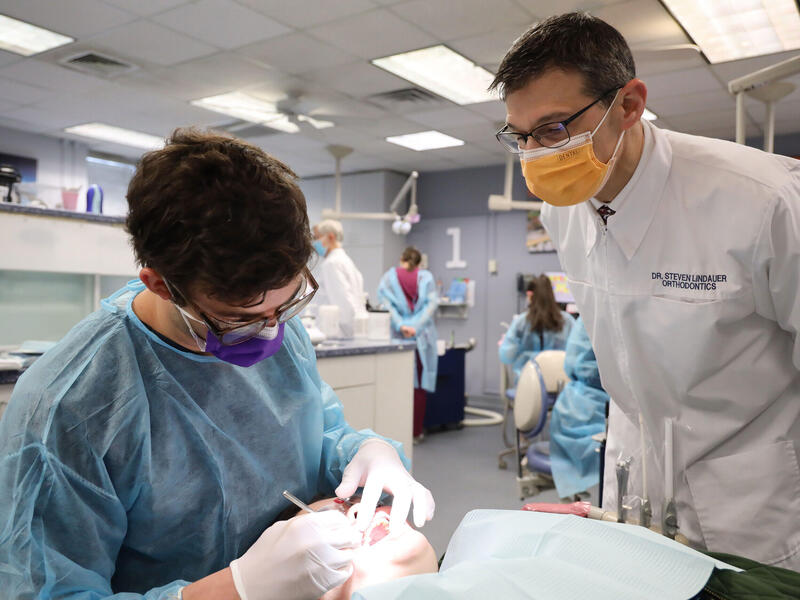 A photo of a dental professor watching a dental student clean a patient's teeth. 