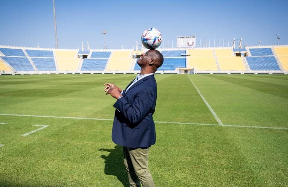 A photo of a man in a suit balancing a socceer ball on his head in the middle of a soccer stadium. 