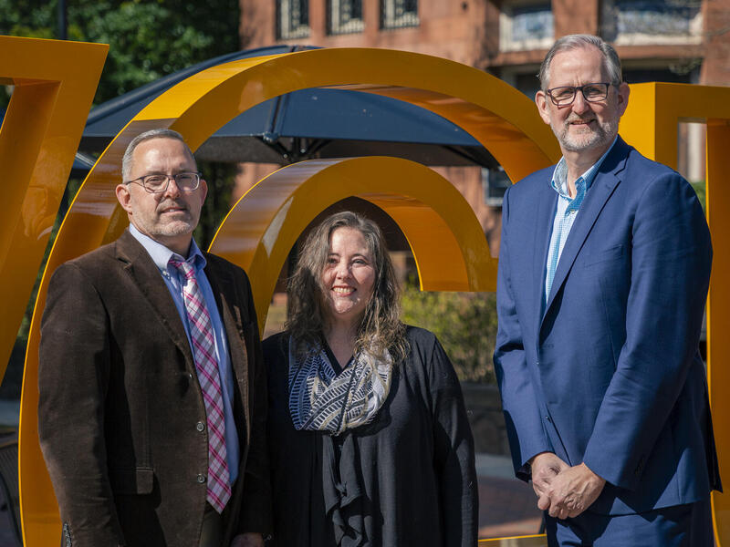 Three people standing in front of a giant letter \"C\"