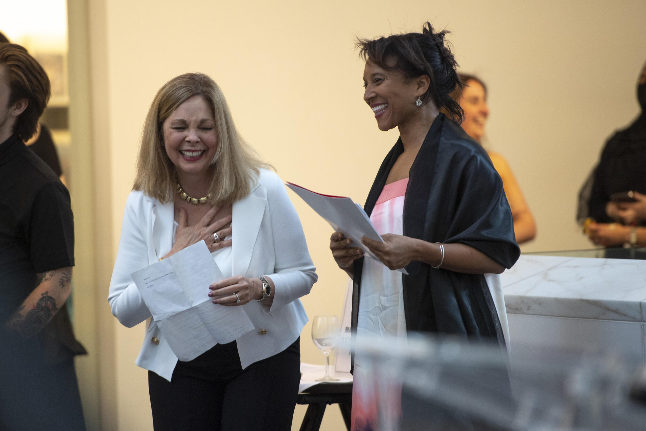 Left to right: Deidra Arrington and Carmenita Higginbotham standing next to each other and holding papers while laughing 