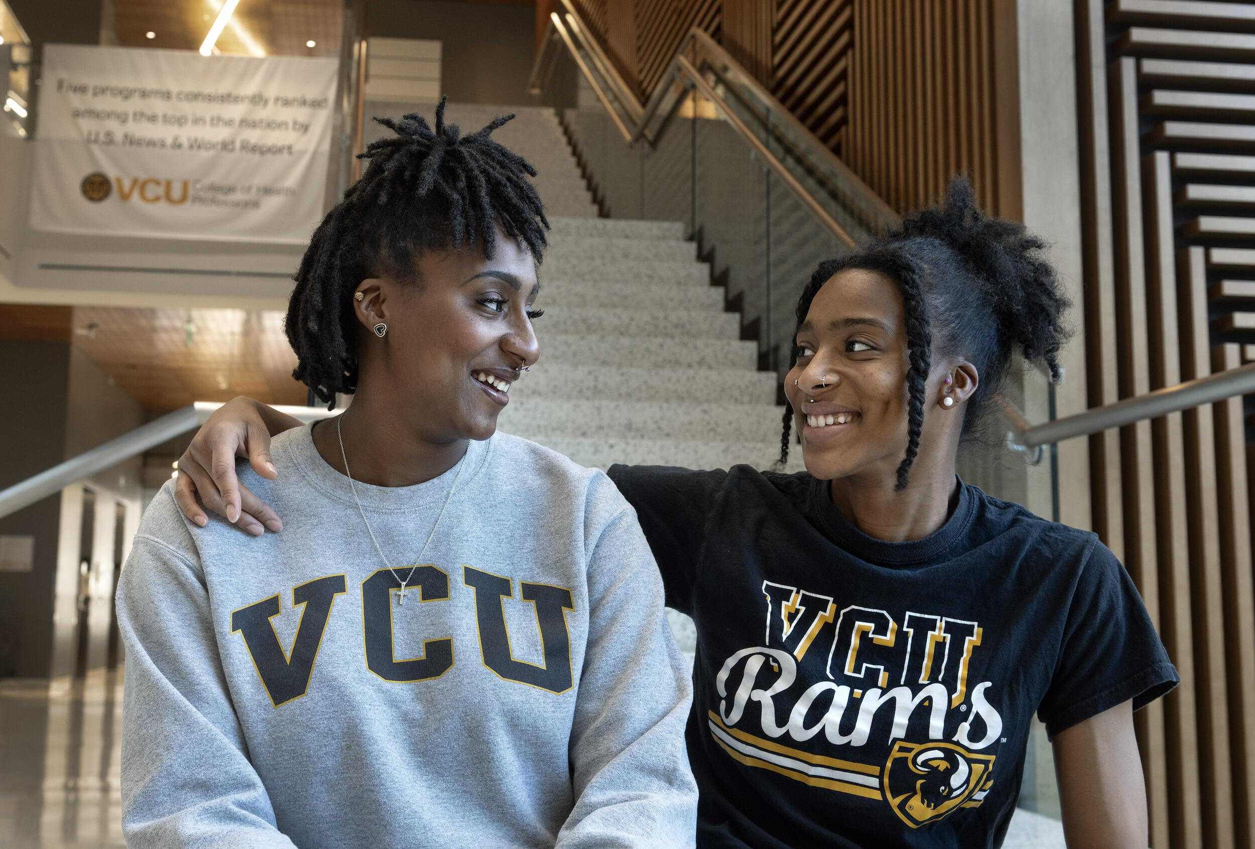 A photo of two women from the chest up. They are sitting on a staircase and have turned their heads to smile at each other. The woman on the right has her left arm draped around the woman on the left's shoulder. The woman on the left is wearing a gray sweatshirt that says \"VCU\" in black letters. The woman on the right is wearing a black T-shirt that says \"VCU Rams\" in yellow and white letters. 