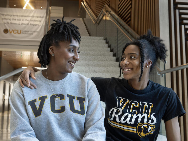 A photo of two women from the chest up. They are sitting on a staircase and have turned their heads to smile at each other. The woman on the right has her left arm draped around the woman on the left's shoulder. The woman on the left is wearing a gray sweatshirt that says \"VCU\" in black letters. The woman on the right is wearing a black T-shirt that says \"VCU Rams\" in yellow and white letters. 