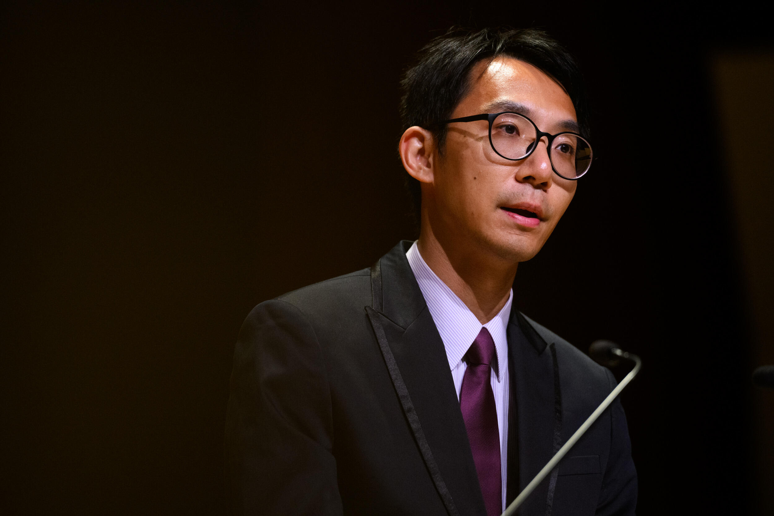 A man in a coat and tie and glass speaks into a microphone.