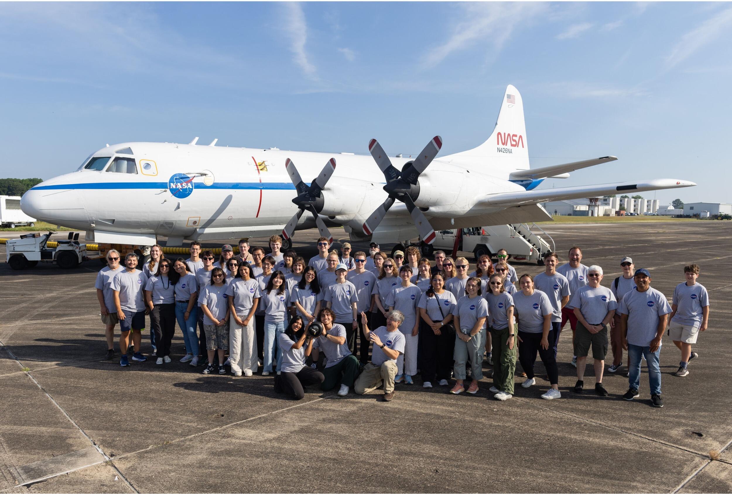 A group photo of a crowd of people standing in front of an airplane which has \"NASA\" written on the side of it. 