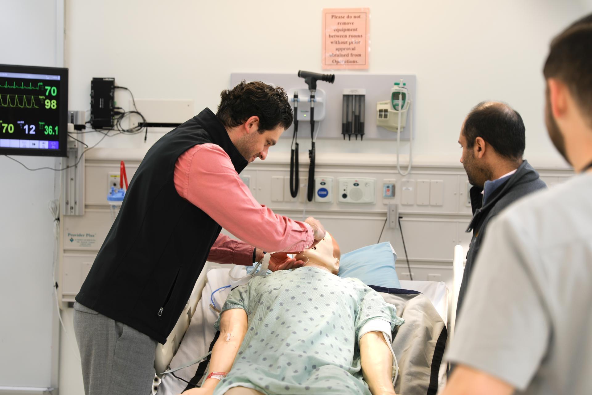A photo of a man examining a manikin wearing scrubs laying on a medical bed. Two men stand to the right and watch the first man examining the manikin. 