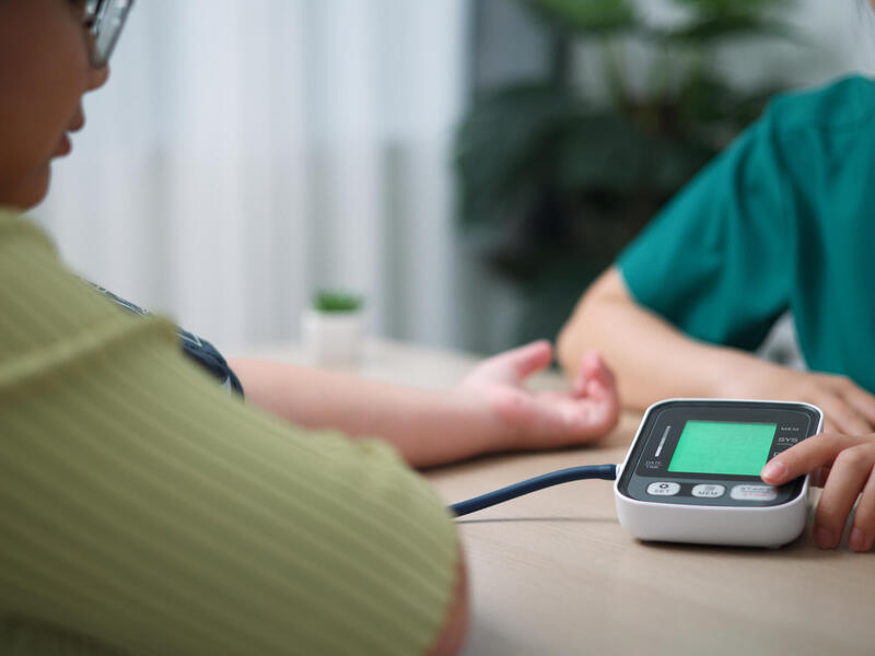 A photo of two people sitting on either side of a table. On the right side of the table is a woman wearing medical scrubs and on the left is a woman wearing a yellow shirt. The woman on the left has a blood pressure monitor on her arm. 